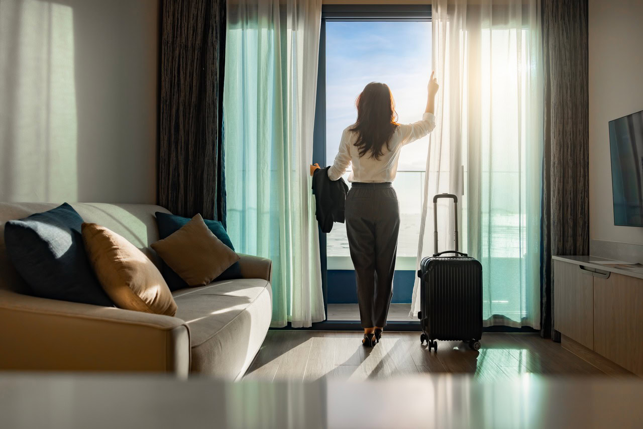 Mulher abrindo a cortina de um hotel com sua mala ao lado para representar a prática de sourcing de hotéis.