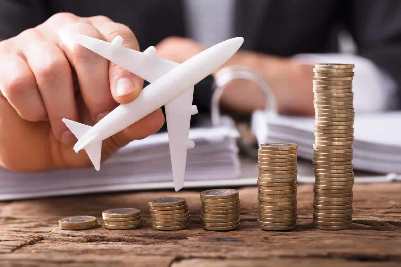 Mão masculina segurando uma miniatura de avião em uma mesa com várias moedas, a fim de calcular o custo de viagem corporativa.