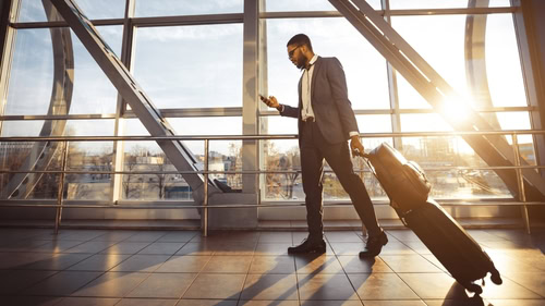 Colaborador no aeroporto com o celular em uma mão e na outra a sua mala tentando descobrir como bater ponto em viagem corporativa.